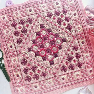 May include: A detailed cross-stitch project in shades of pink and burgundy, featuring a geometric floral design. The needlework is surrounded by embroidery floss, scissors, and decorative buttons. The finished piece is approximately 8 inches square.