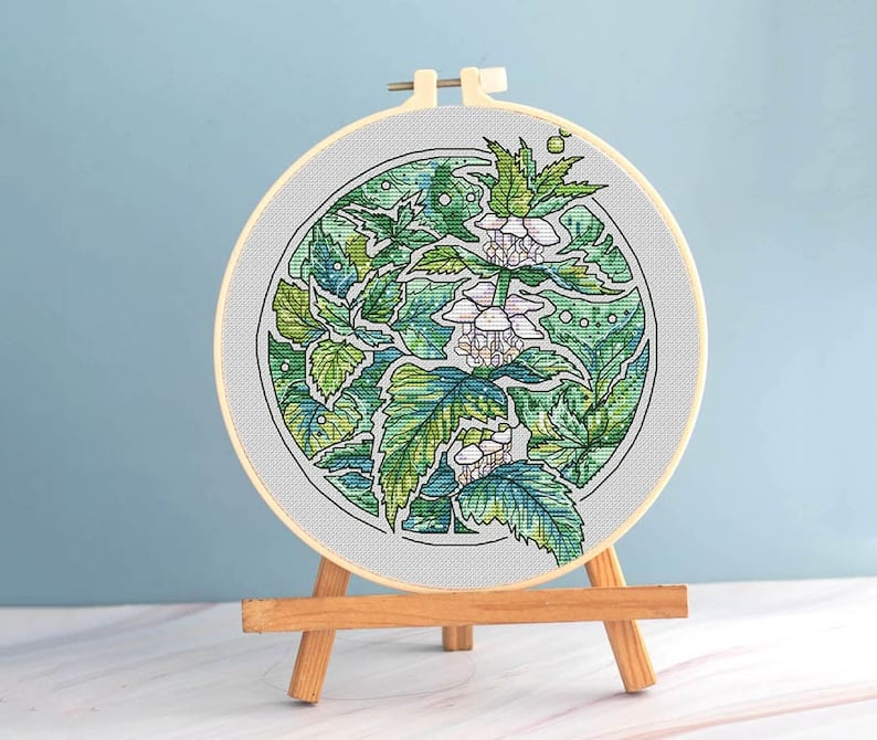 May include: A cross-stitch embroidery hoop featuring a botanical design. The artwork showcases a detailed pattern of green leaves and white flowers within a circular frame. The embroidery is displayed on a small wooden easel.
