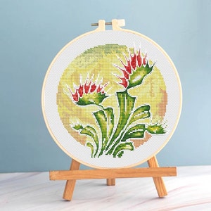 May include: A cross-stitch embroidery of a Venus flytrap plant in a wooden hoop. The plant features red and green details against a yellow and green background. The embroidery is displayed on a small wooden easel.