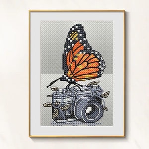 Könnte beinhalten: Kreuzstich-Kunstwerk eines Monarchfalters mit orange-schwarzen Flügeln auf einer grauen Kamera. Die Kamera ist mit Blättern und Ranken verziert. Das Kunstwerk ist in einem goldenen Rahmen gerahmt.
