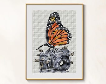 May include: Cross-stitch art of a monarch butterfly with orange and black wings perched on a gray camera. The camera has leaves and vines growing around it. The artwork is framed in a gold frame.