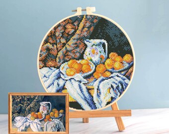Pode incluir: Uma bordado de ponto cruz de uma natureza morta com laranjas, um jarro e um pano listrado branco e azul. O bordado é exibido em um círculo de madeira em um pequeno cavalete de madeira. Uma pintura emoldurada da mesma natureza morta está na frente.