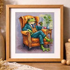 May include: Cross-stitch art of a frog in a suit, smoking a pipe, seated in a tufted armchair. The frog wears a teal suit, a patterned tie, and glasses. A small table holds a vase and teacup. The artwork is framed in a wooden frame.