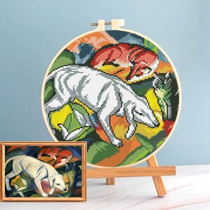 May include: A cross-stitch embroidery hoop depicting a white dog, a red fox, and a black cat, set against a vibrant backdrop of green, orange, and yellow. A framed painting of the same scene is displayed on a small wooden easel.