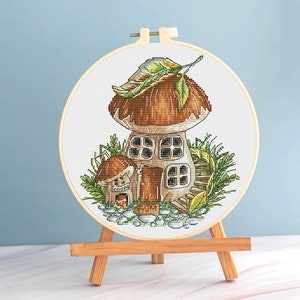 Könnte beinhalten: Ein Kreuzstich-Stickrahmen mit einem skurrilen Pilzhaus-Design. Das Haus hat einen braunen Hut, Fenster und eine Tür, umgeben von grünem Laub und kleinen Steinen. Die Stickerei wird auf einer hölzernen Staffelei präsentiert.