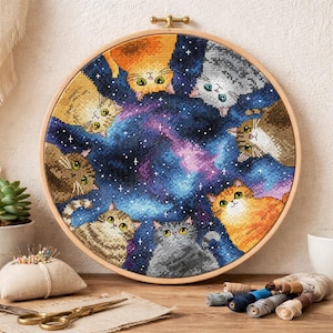 Pode incluir: Um bastidor de bordado em ponto cruz circular com um design celestial com vários gatos contra um fundo estrelado. Os gatos são de várias cores, incluindo laranja, cinza e marrom, com olhos amarelos. O fundo é uma mistura de azul, roxo e rosa.