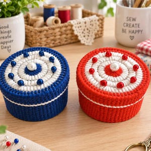 May include: Two round, lidded containers crafted from blue and red yarn, featuring white yarn accents and a central white pearl bead. The lids are decorated with a circular pattern of smaller blue and red beads, creating a textured, handmade appearance.