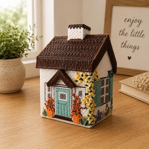 Op de afbeelding: Een handgemaakt miniatuurhuis met een bruin dak, witte muren en een turquoise deur. Het huis is versierd met bloemen in potten en klimmende gele bloemen. De tekst "enjoy the little things" is zichtbaar op de achtergrond.