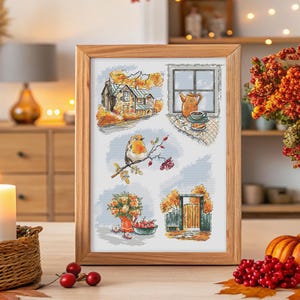 May include: A framed cross-stitch sampler depicting autumnal scenes. The artwork shows a house, a window with a teapot, a bird on a branch, flowers in a vase, and a gate. The frame is made of light-coloured wood.