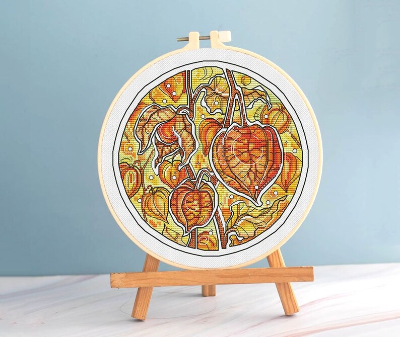 May include: A completed cross-stitch embroidery in a round hoop, featuring a detailed botanical design of orange and yellow Chinese lantern plants. The artwork is displayed on a small wooden easel against a light blue background.