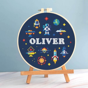 May include: A cross-stitch hoop art piece featuring a navy blue background with space-themed designs. The design includes rockets, planets, and an astronaut, with the name "OLIVER" stitched in white. The hoop is displayed on a small wooden easel.
