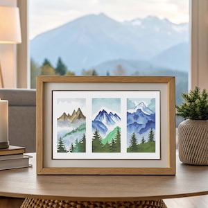 May include: Framed cross-stitch art featuring three mountain landscape panels. The panels depict snow-capped peaks, evergreen trees, and misty valleys in shades of blue, green, and brown. The artwork is displayed in a wooden frame.