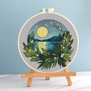 May include: A cross-stitch embroidery hoop featuring a nighttime landscape with a full moon over a body of water. The scene is framed by green and white foliage. The embroidery is displayed on a small wooden easel.