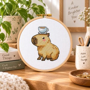 May include: A circular wooden embroidery hoop containing a cross-stitch design of a tan capybara with a small blue and white teacup balanced on its head. The capybara has a dark brown snout, pink cheeks, and is stitched onto white fabric.