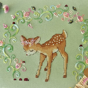 Peut inclure: Broderie au point de croix d'un faon avec une couronne de fleurs, entouré d'une arche de fleurs et de vignes. Le faon est brun avec des taches blanches. La broderie est sur un tissu vert clair. Petites coccinelles et abeilles brodées.