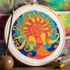 May include: A colorful cross-stitch embroidery in a wooden hoop. The design features a stylized sun with swirling patterns in shades of red, orange, yellow, and blue. An incense stick and a harmonica are in the foreground.