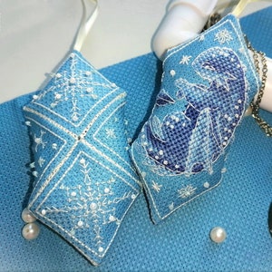 May include: Two light blue, diamond-shaped ornaments with white cross-stitch designs. One features a snowflake, the other a dragon. Both are embellished with white beads and have a cream-colored ribbon for hanging. A winter-themed craft.