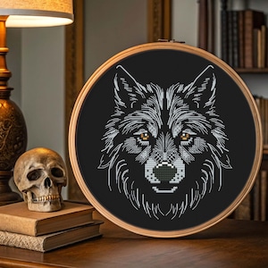 May include: A black cross-stitch embroidery of a wolf's head in a wooden hoop. The wolf has white and gray fur with amber eyes and a black nose. The embroidery is displayed on a wooden surface with a skull and books.