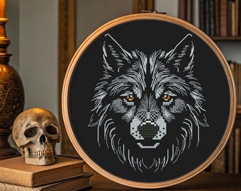 Peut inclure: Broderie au point de croix noire d'une tête de loup dans un cercle en bois. Le loup a une fourrure blanche et grise avec des yeux ambrés et un nez noir. La broderie est exposée sur une surface en bois avec un crâne et des livres.