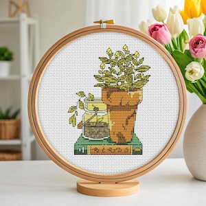 May include: Cross-stitch embroidery of a potted plant, a jar, and a book with the word "BIRDS" on it, displayed in a wooden hoop. The embroidery is set on a white background and displayed on a wooden stand.