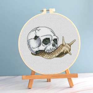 May include: Cross-stitch embroidery of a skull and snail on a white background. The skull is white with gray shading, and the snail is brown and tan. The embroidery is in a wooden hoop, displayed on a small wooden easel.