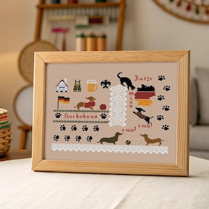 May include: A framed cross-stitch with a beige background, depicting dachshunds, German flag, and other German icons. The design includes the word "Dachshund" and the German word "Wau!" in a decorative font. The frame is made of light-coloured wood.
