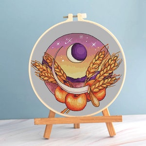 Puede incluir: Bordado de punto de cruz en un aro de madera con un diseño de luna de cosecha. La obra de arte incluye una luna creciente, espigas de trigo y manzanas, sobre un cielo de puesta de sol con estrellas. El bordado se exhibe en un pequeño caballete de madera.