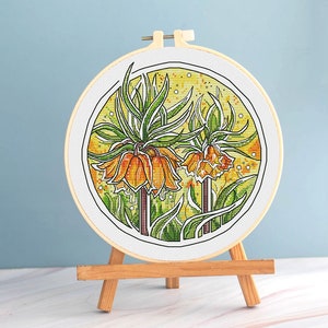 May include: A completed cross-stitch embroidery of orange and yellow flowers with green foliage, set in a round wooden hoop. The artwork is displayed on a small wooden easel against a light blue background.
