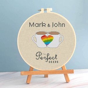 Könnte beinhalten: Ein Kreuzstich-Stickrahmen mit den Namen "Mark & John" und dem Text "The Perfect Blend". Das Design zeigt zwei Kaffeetassen mit einem Regenbogenherz. Der Rahmen wird auf einer kleinen Holzstaffelei präsentiert.