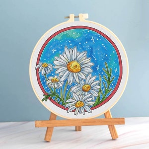 May include: A cross-stitch embroidery hoop featuring a daisy design. The daisies have white petals and yellow centers, set against a blue and green background with star details. The hoop is displayed on a small wooden easel.