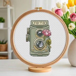 Pode incluir: Bordado em ponto cruz de uma câmera vintage com a palavra "FOTO" no topo. A câmera é verde-oliva com detalhes dourados e uma flor rosa. O bordado está em um bastidor de madeira.