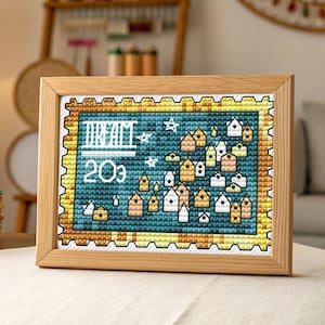May include: A framed cross-stitch artwork featuring a village scene with small houses in various colors. The artwork is set against a teal background with the word "DREAM" and the number "20¢" stitched in white. The frame is made of light-colored wood.
