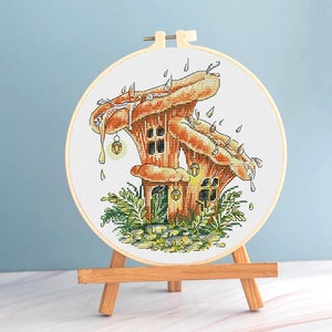 Könnte beinhalten: Kreuzstich-Stickerei eines skurrilen Pilzhauses. Das Design zeigt einen braunen und orangefarbenen Pilz mit Fenstern, tropfenden Details und hängenden Laternen. Die Stickerei ist in einem Holzreifen gefasst und auf einer kleinen Holzstaffelei ausgestellt.