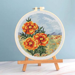 May include: A cross-stitch embroidery of three orange and yellow marigold flowers with green leaves, set against a blue sky and a field of wildflowers. The embroidery is displayed in a round wooden hoop, mounted on a small wooden easel.