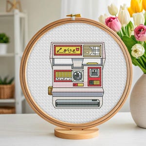 May include: Cross-stitch art of a vintage Polaroid camera, framed in a wooden hoop. The camera is primarily pink, white, and gray, with yellow and red accents. The word "Love" is stitched across the top. The word "Polaroid" is stitched on the front.