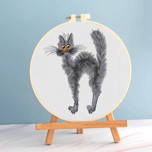 May include: A cross-stitch embroidery hoop featuring a cartoon cat with gray fur, yellow eyes, and a startled expression. The cat is displayed on a small wooden easel against a light blue background. The embroidery hoop is approximately 6 inches in diameter.