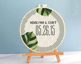 Puede incluir: Un bastidor de bordado de punto de cruz redondo con los nombres "ADALYNN & CURT" y la fecha "05.26.15" en negro. El diseño incluye hojas verdes y un borde a rayas blanco y negro. El bastidor se exhibe en un pequeño caballete de madera.