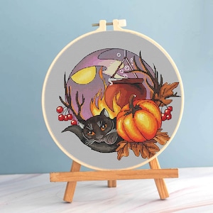 May include: A cross-stitch embroidery in a wooden hoop features a Halloween theme. A black cat with antlers, a pumpkin, and a cauldron are set against a purple and orange background. The embroidery is displayed on a small wooden easel.