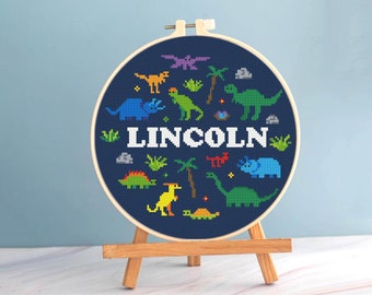 Könnte beinhalten: Ein Kreuzstich-Stickrahmen mit marineblauem Hintergrund und bunten Dinosaurier-Designs. Der Name "LINCOLN" ist in weißen Buchstaben dargestellt. Das Kunstwerk wird auf einer kleinen Holzstaffelei präsentiert.