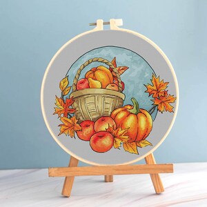 May include: Cross-stitch embroidery in a wooden hoop, featuring a fall harvest scene. A woven basket overflows with pumpkins and apples, surrounded by orange and yellow autumn leaves. The embroidery is displayed on a small wooden easel.