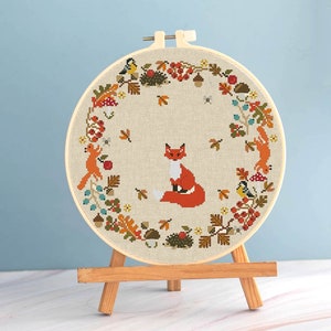 May include: A cross-stitch embroidery hoop with a design of a red fox, surrounded by autumn leaves, berries, squirrels, and birds. The design is on beige fabric, framed by a wooden hoop, and displayed on a small wooden easel.