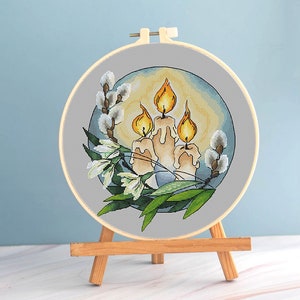 May include: Cross-stitch embroidery in a wooden hoop featuring three lit candles with orange flames. The design includes white flowers, green leaves, and pussy willow branches, set against a light blue background. The embroidery is displayed on a small wooden easel.