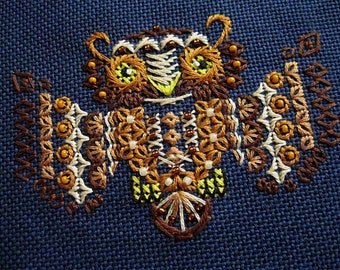 May include: A detailed, embroidered owl design in shades of brown, tan, and yellow, with small bead accents. The owl is centered on a dark blue fabric background. The embroidery features intricate patterns and textures, creating a visually rich and detailed artwork.