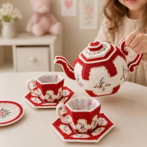 May include: A crocheted tea set featuring a teapot, two cups, two saucers, and a small round mat. Each piece is crafted with white and red yarn, accented with small floral embroidery patterns in pink, blue, and green hues throughout the set.