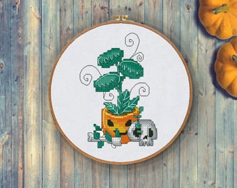 May include: Cross-stitch embroidery in a wooden hoop featuring a Halloween-themed design. The artwork depicts a green plant in a jack-o'-lantern pot, a skull, and a bone. The color palette includes orange, green, white, and gray.