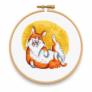 Könnte beinhalten: Eine Kreuzstichstickerei eines braun-weißen Corgi-Hundes mit herausgestreckter Zunge vor einem gelben kreisförmigen Hintergrund. Das Motiv ist in einem runden Holzstickrahmen mit einer Metallschraube zum Festziehen oben eingespannt.