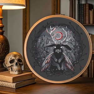 May include: A cross-stitch embroidery in a wooden hoop features a dark gray design of a creature with antlers and moth-like wings. The creature has a red crescent moon above its head. The background includes a forest scene with stars and branches.