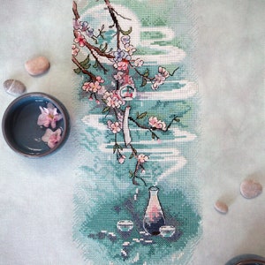 May include: Cross-stitch artwork featuring a Japanese-inspired design. A branch of cherry blossoms with pink flowers and green leaves is depicted above a vase and two cups. The background is a gradient of teal and white, with small stones and bowls of water.