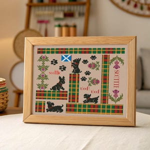 May include: A framed cross-stitch featuring Scottish terriers, paw prints, and thistles. The design includes the text "scottie" and "Woof!" in a tartan-patterned border with a Scottish flag. The frame is made of light-colored wood.