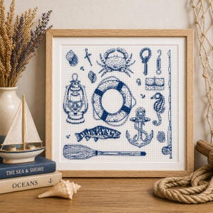 Peut inclure: Une œuvre d'art au point de croix encadrée sur le thème nautique, avec des illustrations bleues et blanches d'objets maritimes. Le motif comprend un crabe, une bouée de sauvetage, une ancre, un hippocampe, un poisson et d'autres éléments nautiques.